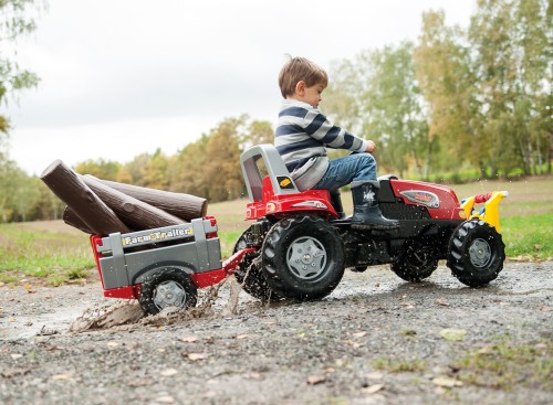 Widok z boku na traktor RollyJunior RT Rolly Toys — kierownica, pedały i przyczepa widoczne podczas jazdy
