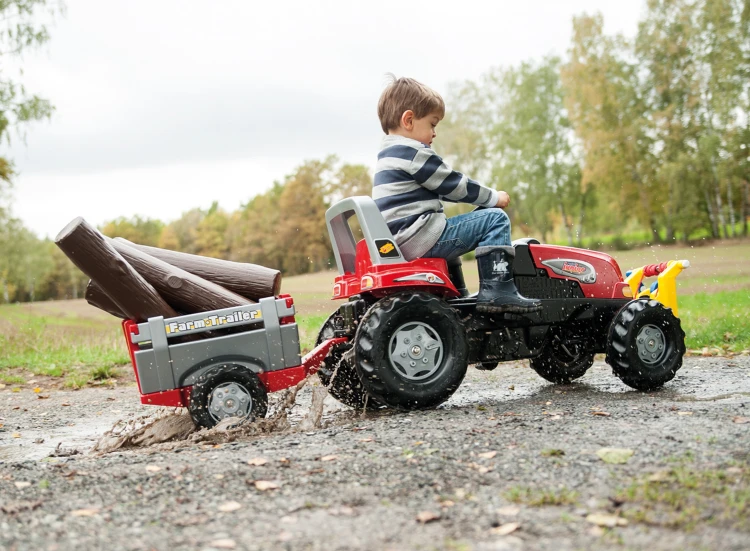 Widok z boku na traktor RollyJunior RT Rolly Toys — kierownica, pedały i przyczepa widoczne podczas jazdy